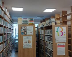 Wnętrze biblioteki wypełnione rzędami wysokich, drewnianych regałów. Po lewej stronie i w głębi półki są ciasno zapełnione książkami, a na ich górnych krawędziach leżą dodatkowe, ułożone poziomo stosy woluminów. Po prawej stronie stoi całkowicie pusty regał przygotowany na książki. Na bocznych ściankach mebli wiszą plakaty informacyjne, w tym logo Miejskiej Biblioteki Publicznej w Przasnyszu oraz ogłoszenie o spotkaniach Klubu Rękodzieła
