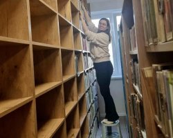 Kobieta w jasnej bluzie i ciemnych spodniach stoi w wąskiej alejce między bibliotecznymi regałami. Aby sięgnąć najwyższych półek drewnianego regału, korzysta z plastikowego stołka (podestu) w kształcie walca. Kobieta uśmiecha się w stronę obiektywu, trzymając dłonie na książkach umieszczonych na samej górze. Po lewej stronie kadru widoczny jest całkowicie pusty regał z kwadratowymi półkami