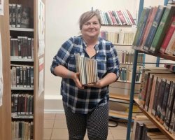 Kobieta w koszuli w niebiesko-białą kratę stoi pośrodku sali bibliotecznej, trzymając oburącz spory pionowy stos książek oparty o brzuch. Kobieta patrzy na obiektyw i uśmiecha się. Wokół niej widoczne są regały z książkami. Na bocznej ściance szafy po lewej stronie wisi plakat zapraszający na „Spotkania Klubu Rękodzieła w każdy wtorek o godzinie 16:00 w Filii nr 1 przy ulicy Orlika 27 w Przasnyszu”