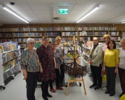 Grupa ośmiu uśmiechniętych kobiet stoi w rzędzie w bibliotece, pozując do zdjęcia wokół gotowego rękodzieła. Na drewnianej sztaludze pośrodku umieszczono okrągłą makramę z brązowych sznurków przypominającą drzewo, którego gałęzie udekorowano papierowymi, kolorowymi listkami. W tle widać białe regały z książkami dziecięcymi i napisami „Poziom I”
