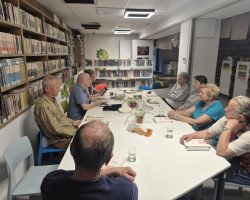 Grupa siedmiu osób – kobiet i mężczyzn – siedzi wokół białego stołu w sali bibliotecznej podczas dyskusji o książkach. Na stole leżą otwarte egzemplarze publikacji, stoją szklanki z wodą oraz talerzyki z drobnymi przekąskami. Po lewej stronie widoczne są wysokie, drewniane regały wypełnione książkami