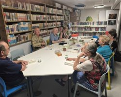 Uczestnicy spotkania czytelniczego siedzą przy podłużnym, białym stole w bibliotece. Zdjęcie wykonano z perspektywy ukazującej tył głowy jednego z mężczyzn na pierwszym planie. Pozostałe osoby słuchają wypowiedzi lub przyglądają się książkom leżącym na stole. W tle widać regały z literaturą oraz rysunek zielonego smoka na dalekiej ścianie