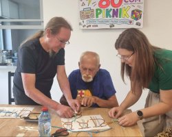 Trzy osoby – mężczyzna w czarnej koszulce, starszy mężczyzna w niebieskiej koszulce oraz kobieta w zielonej bluzce – pochylają się nad drewnianym stołem. Wspólnie pracują nad skomplikowanym splotem sznurka, korzystając ze schematów wydrukowanych na kartkach A4. Na ścianie w tle wiszą plakaty promocyjne