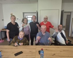 Siedmiu uczestników zajęć rękodzielniczych – kobiety i mężczyźni (w tym osoba na wózku inwalidzkim) – pozuje razem do pamiątkowej fotografii. Każdy z dumą trzyma w dłoni i prezentuje ukończony kolorowy brelok ze sznurka