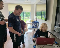Kobieta o blond włosach siedzi przy stanowisku komputerowym i obsługuje myszką czerwonego laptopa. Obok niej stoją trzej uczestnicy spotkania, w tym mężczyzna w ciemnej koszulce, który z uśmiechem przygląda się pracy na komputerze