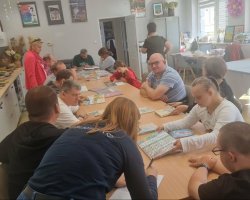 Szerokie ujęcie z góry na grupę kilkunastu osób siedzących przy długim, drewnianym stole w jasnej świetlicy. Uczestnicy, w tym osoby dorosłe i młodzież, przeglądają rozłożone na stole książki i ilustracje. Na pierwszym planie widać kobietę w ciemnym ubraniu. W tle widać białe szafki z wystawą rękodzieła (ceramika, suche bukiety) oraz plakaty na ścianach. Panuje swobodna atmosfera skupienia