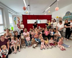 Pamiątkowe, grupowe zdjęcie w jasnym pomieszczeniu bibliotecznym lub przedszkolnym. Kilkadziesięcioro małych dzieci (w wieku żłobkowym i przedszkolnym) siedzi lub stoi w towarzystwie kilku dorosłych kobiet. W tle widoczna jest czerwona, materiałowa kurtyna, lampiony pod sufitem oraz baner z logo Miejskiej Biblioteki Publicznej