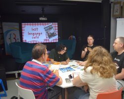 Pięcioro uczestników siedzi wokół białego stołu, grając w planszową grę językową. Na ekranie projektora w tle wyświetla się plansza z napisem "last language café". Na stole, oprócz elementów gry, leżą telefony komórkowe oraz miseczki z przekąskami. Uczestnicy są skupieni, rozmawiają i wskazują karty na planszy
