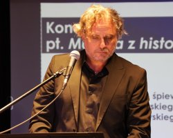 Bliskie ujęcie mężczyzny z kręconymi włosami, w ciemnym garniturze i czarnej koszuli, stojącego przy mikrofonie ze spuszczonym wzrokiem. W tle widać fragment slajdu z tekstem „Koncert pt. Muzyka z historią w tle”