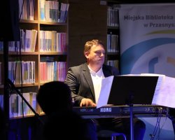 Mężczyzna w garniturze gra na instrumentach klawiszowych Korg umieszczonych na scenie. Obok niego stoi regał pełen książek oraz roll-up Miejskiej Biblioteki Publicznej w Przasnyszu