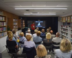 Szerokie ujęcie sali bibliotecznej z perspektywy tyłu widowni. Daniel Kocuj stoi na tle czarnej ściany obok dużego ekranu wyświetlającego napis „Bike’owa podróż z Sydney do Szczecina” z grafiką rowerzysty. Autor, ubrany w czerwoną koszulkę z godłem Polski, przemawia do zgromadzonej w rzędach krzeseł publiczności. Z obu stron salę otaczają wysokie regały pełne książek