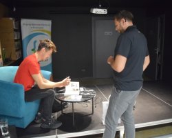 Daniel Kocuj siedzi w turkusowym fotelu na niskim podwyższeniu i podpisuje egzemplarz książki leżący na okrągłym, czarnym stoliku. Autor ma na sobie czerwoną koszulkę, ciemne spodnie i sportowe buty. Przed podwyższeniem stoi odwrócony tyłem mężczyzna w ciemnej koszulce polo i szarych spodniach, który czeka na autograf. W tle widoczny jest baner z logo Miejskiej Biblioteki Publicznej w Przasnyszu