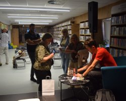 Daniel Kocuj w czerwonej koszulce siedzi w turkusowym fotelu i podpisuje książki przy małym, okrągłym stoliku w bibliotece. Przed nim stoi grupa czytelników – na pierwszym planie kobieta w kwiecistej bluzce przegląda coś w telefonie, obok stoi mężczyzna z ramionami splecionymi na piersi, a w tle czekają kolejne dwie kobiety. W głębi sali widać rzędy pustych krzeseł i regały z książkami