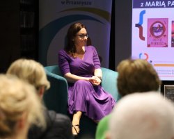Maria Paszyńska w fioletowej sukience i okularach siedzi w turkusowym fotelu podczas spotkania autorskiego. Pisarka uśmiecha się, patrząc w stronę publiczności, której zarysy postaci widać na pierwszym planie