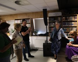 Maria Paszyńska podpisuje książkę przy stoliku w bibliotece. Obok niej stoi kobieta w jeansowej kurtce, a po lewej stronie w kolejce czekają inni uczestnicy spotkania, w tym mężczyzna robiący zdjęcie smartfonem. W tle regały pełne książek