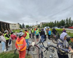 Duża grupa rowerzystów w różnym wieku zebrała się na wybrukowanym placu obok drewnianej tężni solankowej. Wielu uczestników ma na sobie odblaskowe kamizelki oraz kaski ochronne. W tle widoczne są zaparkowane samochody, miejska zabudowa oraz pochmurne niebo
