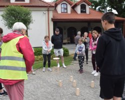 Uczestnicy pikniku biorą udział w zabawie integracyjnej na wybrukowanym dziedzińcu przed budynkiem o dachu z czerwonej dachówki. Trzymając w dłoniach naprężone sznurki, wspólnie sterują metalowym chwytakiem zawieszonym na środku, próbując ustawić pionowo drewniane klocki na ziemi