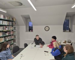 Uczestnicy spotkania w bibliotece na poddaszu, sfotografowani pod innym kątem. Mężczyzna siedzący na środku przegląda leżące przed nim dokumenty, gestykulując dłonią. Cztery kobiety siedzące wokół stołu słuchają go. Po prawej stronie widać regał pełen książek, a w głębi stolik z czajnikiem elektrycznym