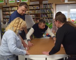 Grupa pięciu osób współpracuje przy białym stole w jasnej sali bibliotecznej. Wspólnie dociskają i wyrównują duży arkusz brązowej tektury. W tle widoczne są regały z książkami oraz zawieszone pod sufitem dziecięce rysunki o tematyce jesiennej