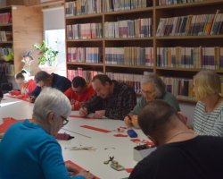Bliskie ujęcie na osoby siedzące przy stole przed regałami z książkami. Uczestnicy za pomocą ołówków i linijek odmierzają i odrysowują linie na czerwonych arkuszach papieru, przygotowując elementy do wycięcia