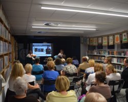 Szerokie ujęcie z głębi sali bibliotecznej na spotkanie autorskie z pisarzem Przemysławem Piotrowskim. Pisarz i moderatorka siedzą na scenie na niebieskich fotelach, a przed nimi zgromadzona publiczność siedząca w rzędach krzeseł. Regały z książkami otaczają salę z obu stron, a na ścianach wiszą rysunki. Na ekranie w tle widoczny slajd tytułowy