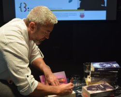 Zbliżenie z profilu na pisarza Przemysława Piotrowskiego, ubranego w białą koszulę, który pochyla się i podpisuje książkę dla czytelnika. Na stoliku obok niego leży stos jego książek, w tym widoczne tytuły 'MARXIST', 'FETYSZ', 'ANUBIS', 'SMOLARZ' i 'BAGNO'. Z boku widać fragmenty dłoni innej osoby