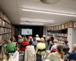 Perspektywa z boku sali bibliotecznej, ukazująca liczną grupę dorosłych osób słuchających wykładu. Nad regałami po prawej stronie wiszą oprawione rysunki dziecięce. Na pierwszym planie po lewej stronie stoi stolik z cateringiem, na którym umieszczono duży, metalowy termos gastronomiczny na napoje oraz białe filiżanki