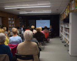 Widok z tyłu na salę biblioteczną wypełnioną siedzącymi na krzesłach uczestnikami wykładu. W głębi sali, na środku czarnej ściany, wyświetlany jest jasny slajd prezentacji z napisem „Jak odzyskać siebie i żyć bez lęku”. Po bokach ekranu siedzą prowadzące. Wokół sali ciągną się regały z książkami
