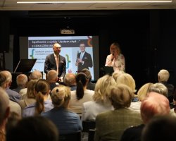 Mężczyzna w garniturze siedzi na scenie z mikrofonem w dłoni, a obok niego stoi kobieta przemawiająca do mikrofonu. Przed sceną siedzi zgromadzona publiczność. W tle ekran z plakatem wydarzenia „Jestem stąd…”