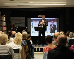 Łukasz Motyka gra na klarnecie na podwyższeniu, w tle widoczna pianistka oraz ekran projekcyjny. Na pierwszym planie tyłem do obiektywu siedzi publiczność zapełniająca salę biblioteczną