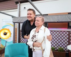 Robert Kudelski pozuje do pamiątkowej fotografii z kobietą na scenie po wydarzeniu. Artysta w czarnej koszuli obejmuje ramieniem uśmiechniętą uczestniczkę, która ma na sobie białą marynarkę, bluzkę we wzory i jasne spodnie. W tle widać dekoracyjną, złotą lampę oraz drewniane kratki na ścianie budynku