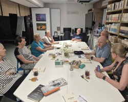 Dyskusja grupy siedmiu czytelników siedzących przy podłużnym, białym stole w nowoczesnej przestrzeni bibliotecznej. Na blacie wyeksponowane są okładki różnych książek przygotowanych do rozmowy, a także filiżanki z napojami. Uczestnicy spotkania są skupieni i wymieniają opinie. Po prawej stronie widoczne są wysokie, drewniane regały wypełnione książkami
