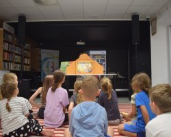 Dzieci siedzą tyłem na pasiastym dywanie, oglądając przedstawienie prezentowane w drewnianej skrzynce teatrzyku Kamishibai, która stoi na stole. W tle widać regał z książkami i kolorowe koło fortuny