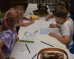 Grupa dzieci siedzi przy podłużnym, białym stole i rysuje kredkami na białych kartkach papieru. Na stole leżą materiały edukacyjne oraz wiklinowe koszyki z przyborami plastycznymi
