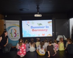 Grupa dzieci siedzi tyłem na podłodze, oglądając wyświetlany na dużym ekranie slajd z napisem „Summer in Germany” i rysunkiem żółwia morskiego. Po lewej stronie na podwyższeniu siedzi prowadząca zajęcia, obok której stoi pionowy baner reklamowy biblioteki