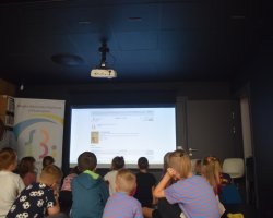 Grupa dzieci siedzi tyłem na podłodze w zaciemnionej sali, oglądając wyświetlaną na dużym ekranie stronę internetową bibliotecznej wyszukiwarki książek. Obok ekranu stoi baner reklamowy, a z boku przygląda się im opiekunka