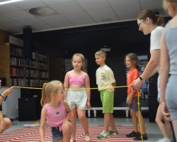 Dzieci i opiekunowie biorą udział w zabawie ruchowej w sali bibliotecznej. Dwie dorosłe osoby trzymają napięty żółty sznur, pod którym przechodzą dzieci stojące w kolejce na dywanie w biało-czerwone pasy. W tle widać regały z książkami