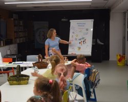 Kobieta stoi przy tablicy flipchart z napisem „Bezpieczne wakacje” i wskazuje na przypięte do niej rysunki edukacyjne. Przed nią, przy białym stole, siedzi tyłem grupa dzieci