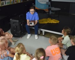 Kobieta w okularach i niebieskiej koszulce w paski siedzi na podwyższeniu, pokazując mały, retro projektor grupie dzieci siedzących przed nią w półkolu na podłodze