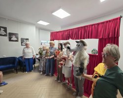 Kobiety stojące przed czerwoną kurtyną teatralną prezentują lalki i pacynki, w tym postać wilka, gajowego oraz babci. Na pierwszym planie po prawej stronie uśmiecha się kobieta w zielonej bluzce trzymająca wełnianą pacynkę wilka