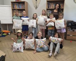 Grupa dziewięciorga uśmiechniętych dzieci oraz stojąca z tyłu ciemnowłosa instruktorka pozują w przytulnej sali bibliotecznej, prezentując białe, ozdobione kolorowymi rysunkami poduszki. W tle widoczne są regały wypełnione książkami, flipchart oraz drukarka