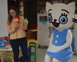 Maskotka Kici Koci siedzi na wysokim krześle w bibliotece. Obok niej stoi bibliotekarka, która trzyma w rękach kolorowe rekwizyty w kształcie głowy kota na patyczkach. Na małym stoliku obok leżą książeczki z serii o Kici Koci