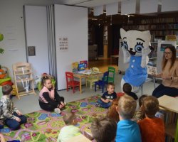 Grupa dzieci siedzi na kolorowym dywanie w kąciku dziecięcym i słucha bajki. Bibliotekarka siedzi obok maskotki Kici Koci i czyta na głos książeczkę. Maskotka Kicia Kocia przykłada ręce do policzków w geście zdziwienia, animując czytaną historię