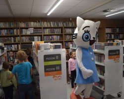 Spacer między regałami. Maskotka Kicia Kocia idzie korytarzem bibliotecznym, a za nią i obok niej między białymi regałami przemykają dzieci. Widoczne są oznaczenia poziomów książek dla dzieci