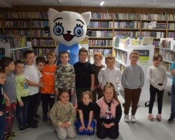 Pamiątkowe zdjęcie grupy dzieci z maskotką Kici Koci w bibliotece. Wielka maskotka w niebieskiej sukience stoi w centrum, otoczona uśmiechniętymi dziećmi. Część dzieci stoi, a kilka dziewczynek klęczy w pierwszym rzędzie na tle regałów wypełnionych książkami