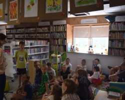 Uczestnicy warsztatów siedzą na kolorowym dywanie i pufach w strefie dla dzieci w bibliotece. Po lewej stronie stoi uśmiechnięta prowadząca, która rozmawia z jednym z chłopców. Nad regałami z książkami wiszą dziecięce rysunki, a w głębi znajduje się jasne okno z zabawkami na parapecie