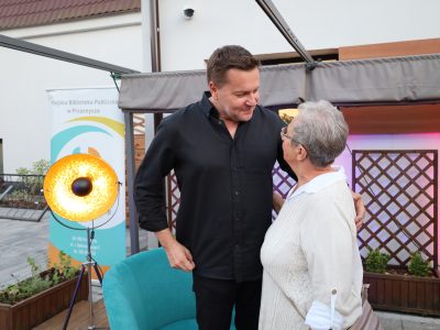 Robert Kudelski rozmawia z bliska ze starszą kobietą na scenie. Artysta nachyla się z ciepłym uśmiechem w stronę uczestniczki spotkania, trzymając dłoń na jej ramieniu. Kobieta ma krótkie, siwe włosy, nosi okulary i jasną, ażurową bluzkę. W tle stoi złota lampa sceniczna z włączonym światłem