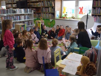 Grupa dzieci siedzi w kręgu na kolorowym dywanie w bibliotece, uważnie słuchając bibliotekarki. Jedno dziecko w środku podnosi rękę. W tle widać regały z książkami oraz okno ozdobione papierowymi zającami. Na pierwszym planie znajduje się stolik z kolorowankami