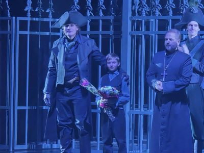 Zbliżenie na scenę w niebieskim oświetleniu. Aktor w płaszczu i dwurożnym kapeluszu trzyma dłoń na ramieniu chłopca z kwiatami, obok nich stoi aktor w stroju duchownego