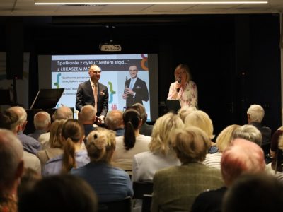 Mężczyzna w garniturze siedzi na scenie z mikrofonem w dłoni, a obok niego stoi kobieta przemawiająca do mikrofonu. Przed sceną siedzi zgromadzona publiczność. W tle ekran z plakatem wydarzenia „Jestem stąd…”